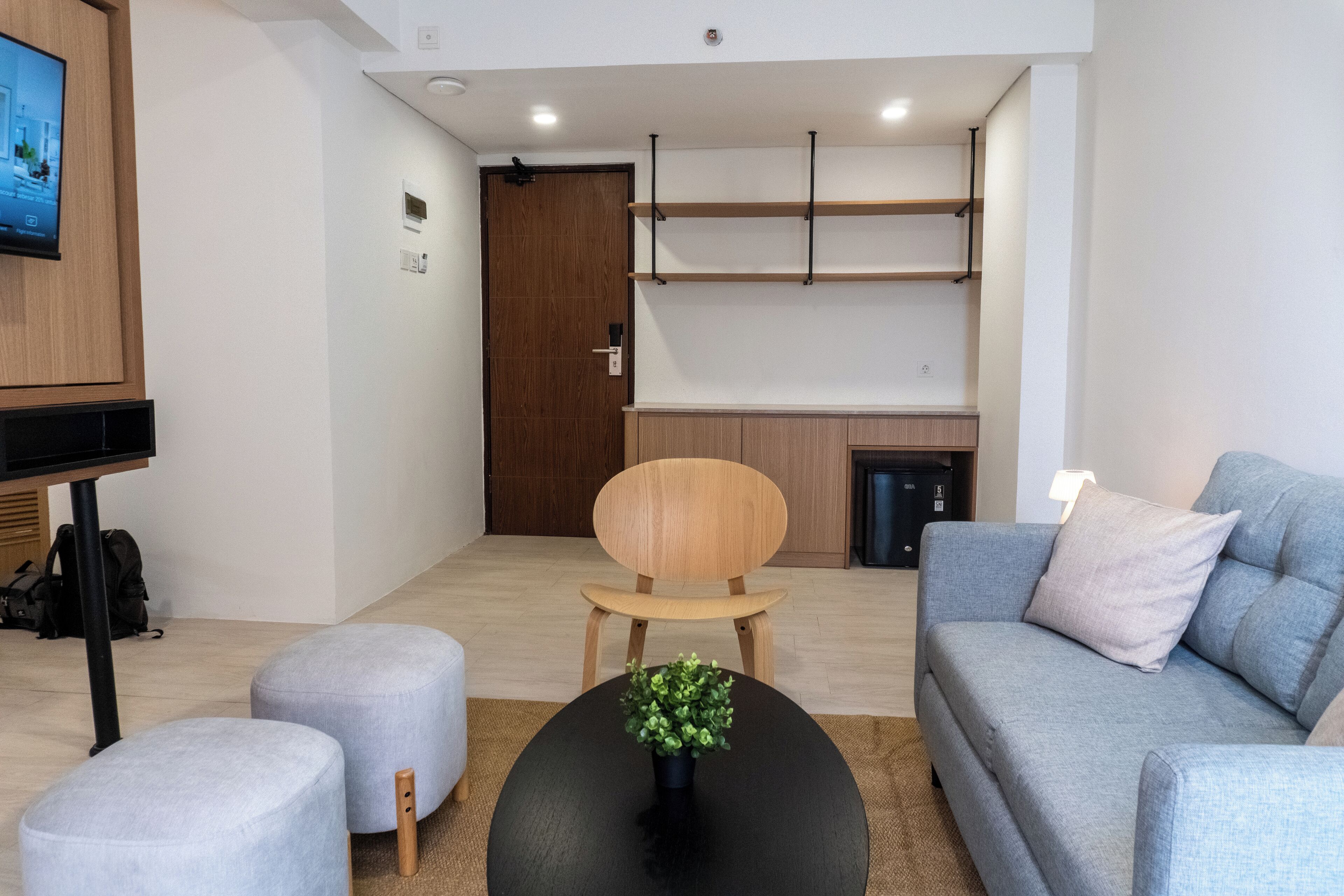 A modern living room with a grey sofa, a wooden chair, and a black coffee table with a plant. There is a TV mounted on the wall, a wooden cabinet, and a kitchen area in the background.