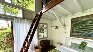 A bedroom with a wooden ladder leading to a balcony, a bed with patterned bedding, a mirror, and a large painting on the wall.