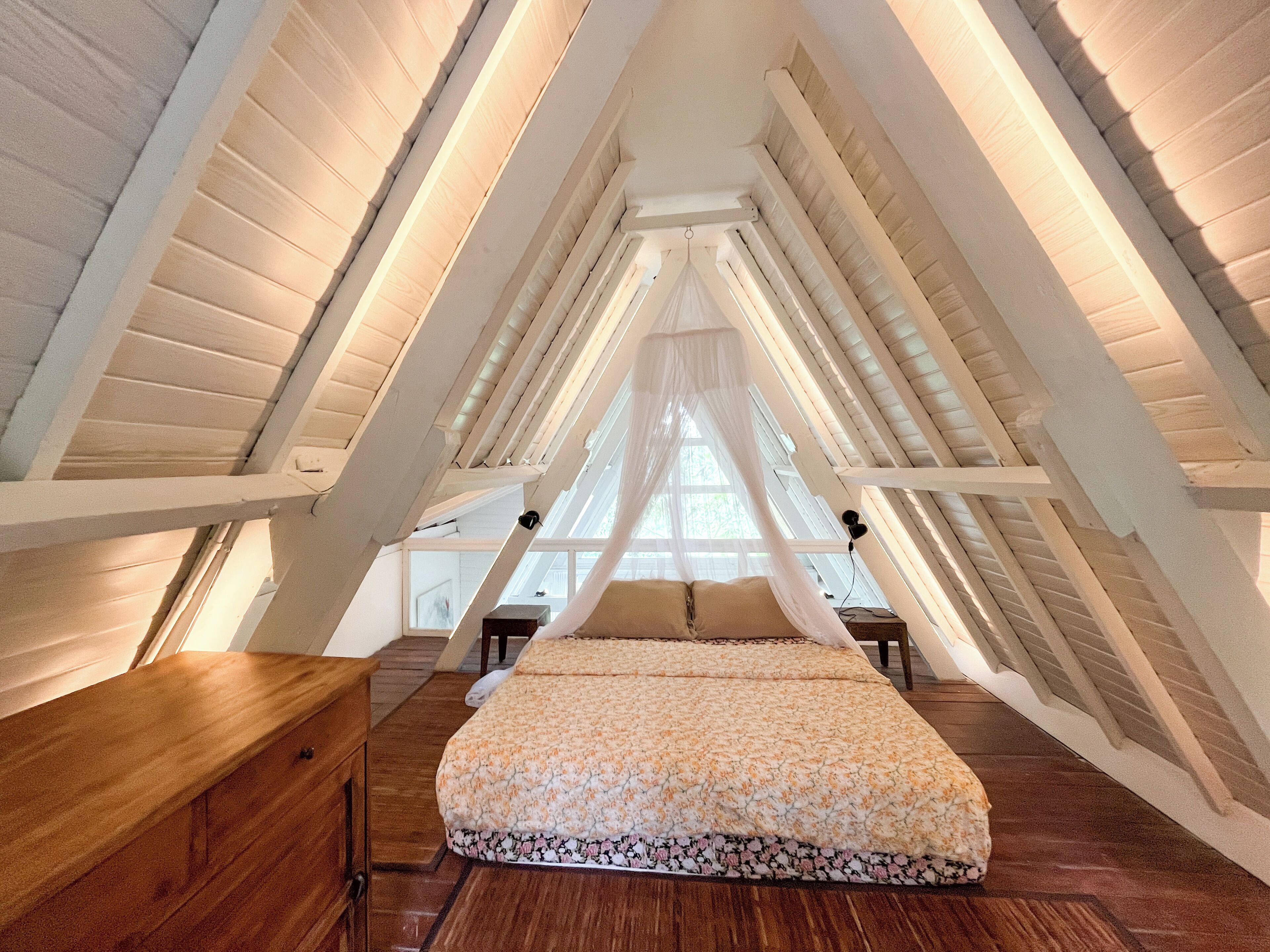 A bedroom with a sloped ceiling, a bed with a floral bedspread, a wooden dresser, and a window with a sheer curtain.