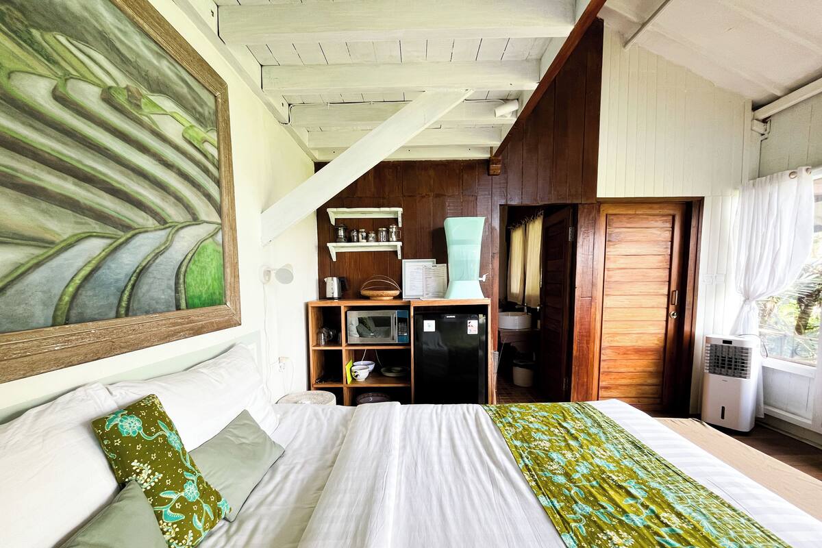 Un dormitorio con cama, repisa de madera con utensilios de cocina, microondas y ventana con cortinas.