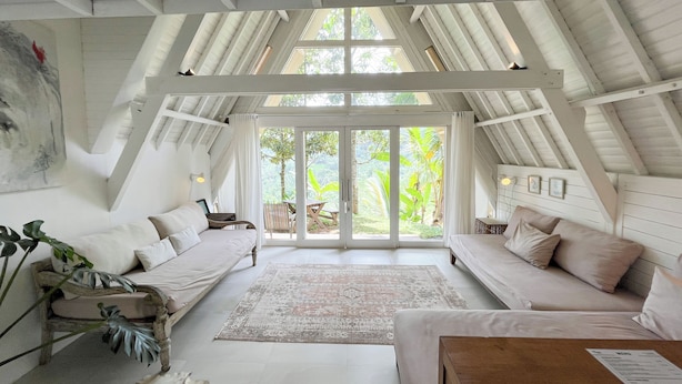 A spacious room with a high ceiling and exposed beams, featuring a large sliding glass door leading to a lush garden.