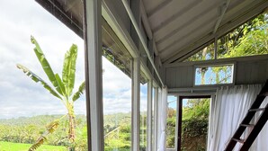 A room with large windows overlooking a lush garden, a wooden chair, a round table, and a staircase leading outside.