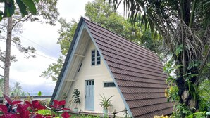A small, single-story house with a sloped roof, surrounded by lush greenery and various plants.