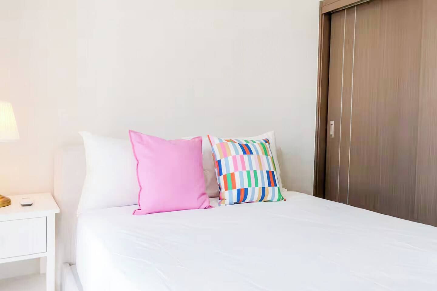 Quarto de hotel com cama, mesa de cabeceira, luminária e porta de madeira.