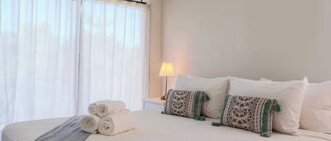 Une chambre d’hôtel avec un grand lit, du linge de lit blanc, des serviettes pliées, des coussins à motifs, une lampe de chevet et une fenêtre avec des rideaux légers.