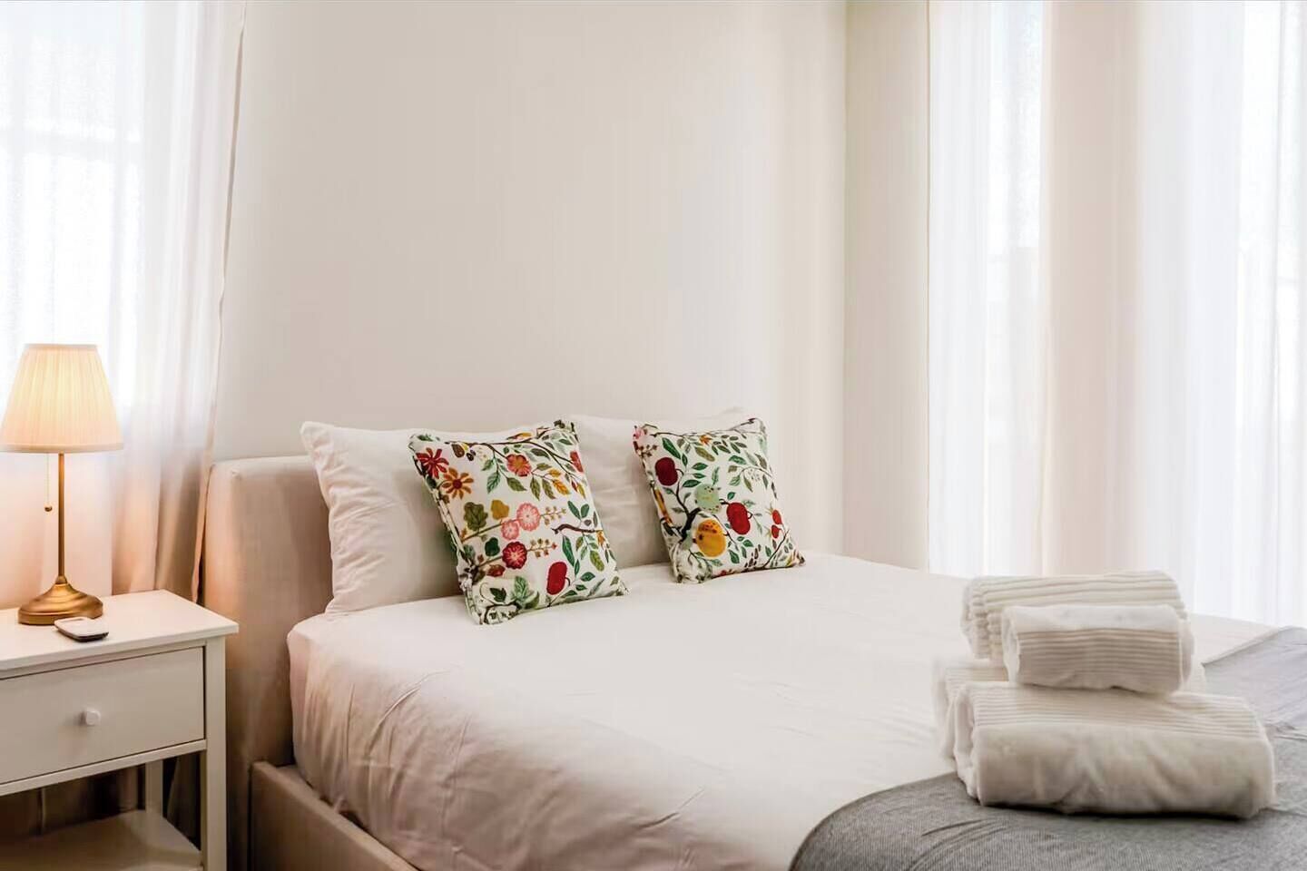 Een hotelkamer met een bed, twee sierkussens met bloemenprint, een bedlampje, en een netjes opgemaakt bed met gevouwen handdoeken.