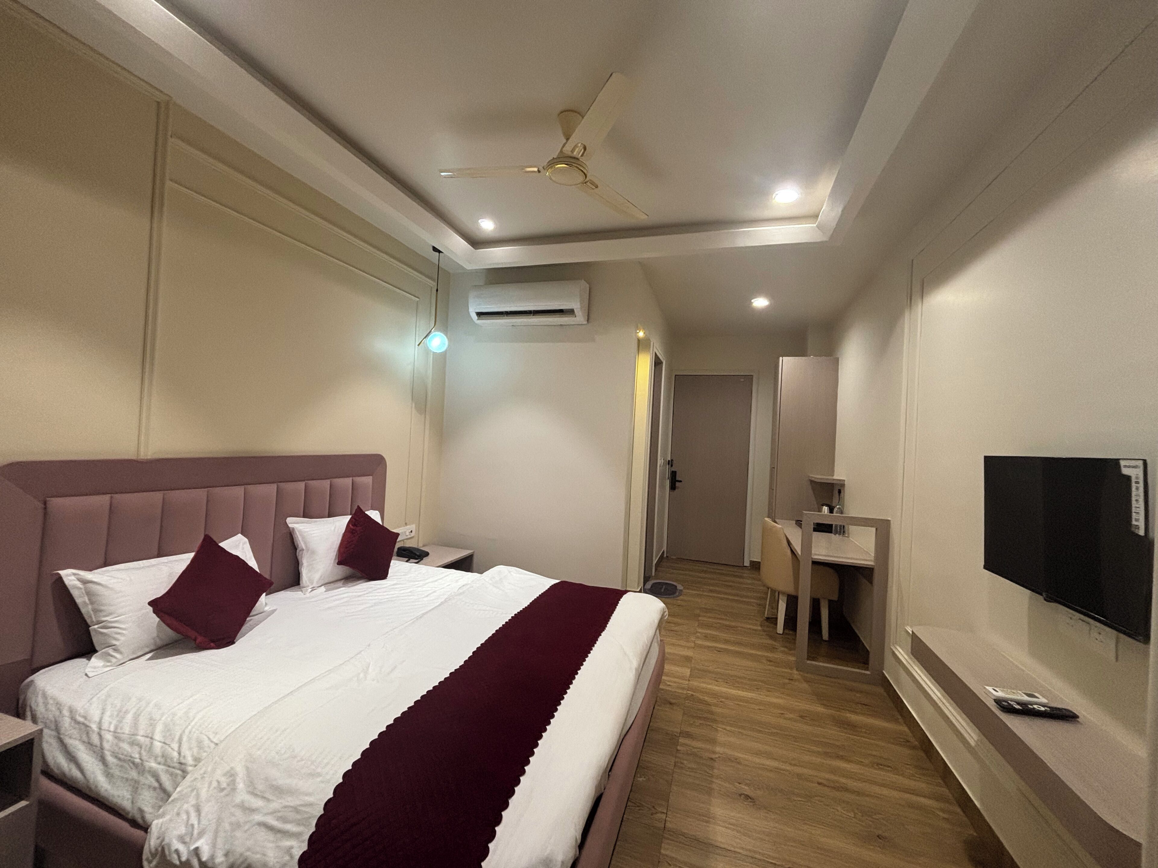 A hotel room with a large bed, a desk, a television, and an air conditioning unit.