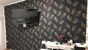 Een kamer met behangpapier met een bladmotief, een flatscreen televisie aan de muur, een witte tafel met vier stoelen, en een vaas met roze bloemen.