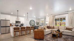 Un salon moderne avec un canapé, un pouf et une table basse. On y trouve également un coin cuisine avec des meubles blancs et un réfrigérateur. Un coin repas avec des chaises et une table est visible.