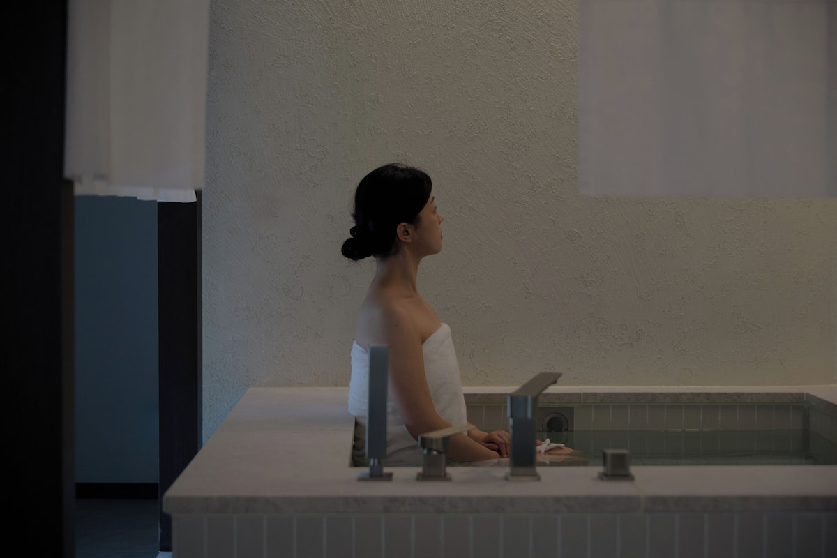 Una persona sentada junto a una bañera en un baño con pared lisa y piso de baldosas.