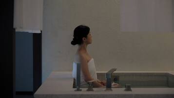 A person sitting by a bathtub in a bathroom with a plain wall and a tiled floor.
