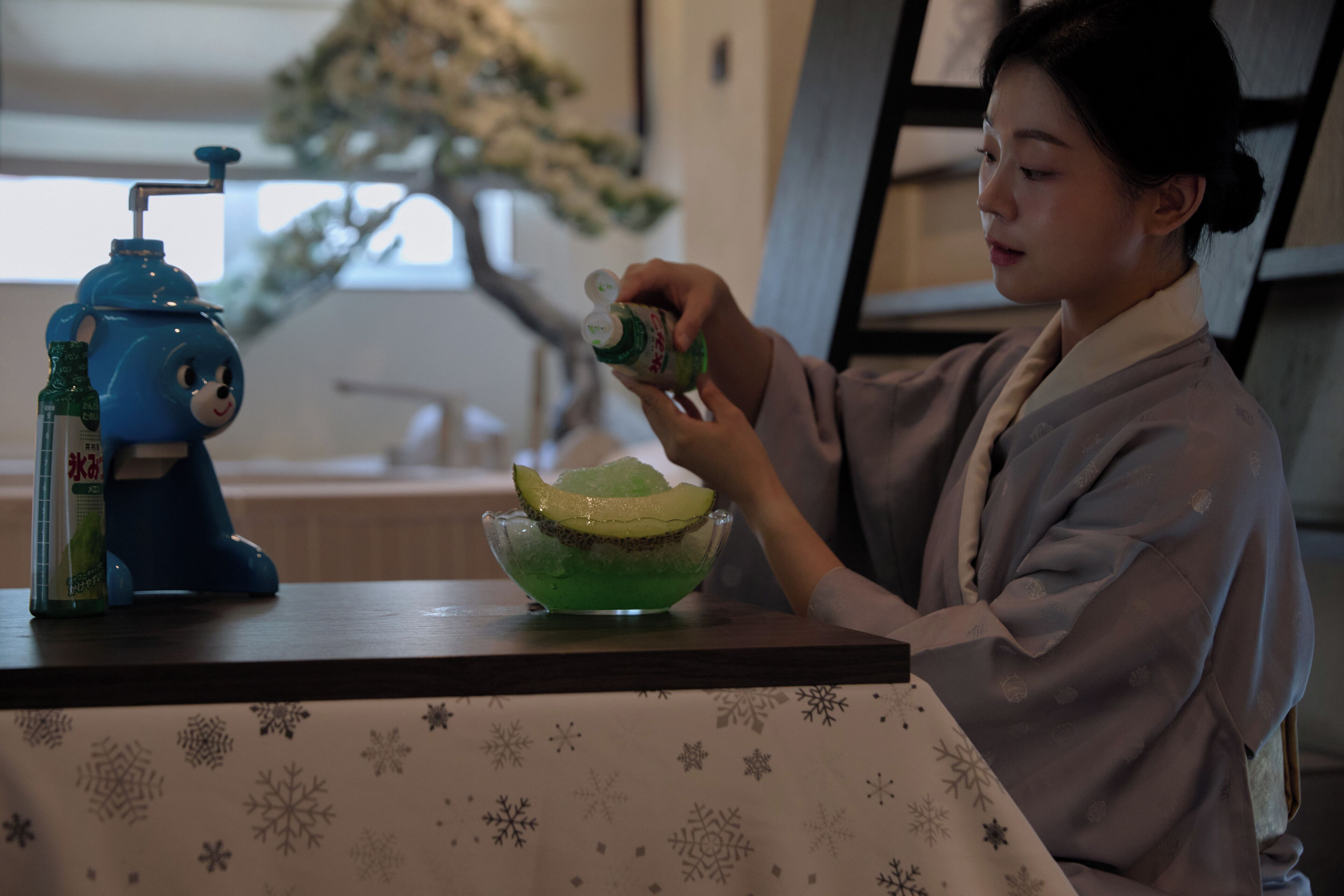 A person in traditional attire preparing a drink with a blue dispenser and a bowl on a table with a snowflake pattern.