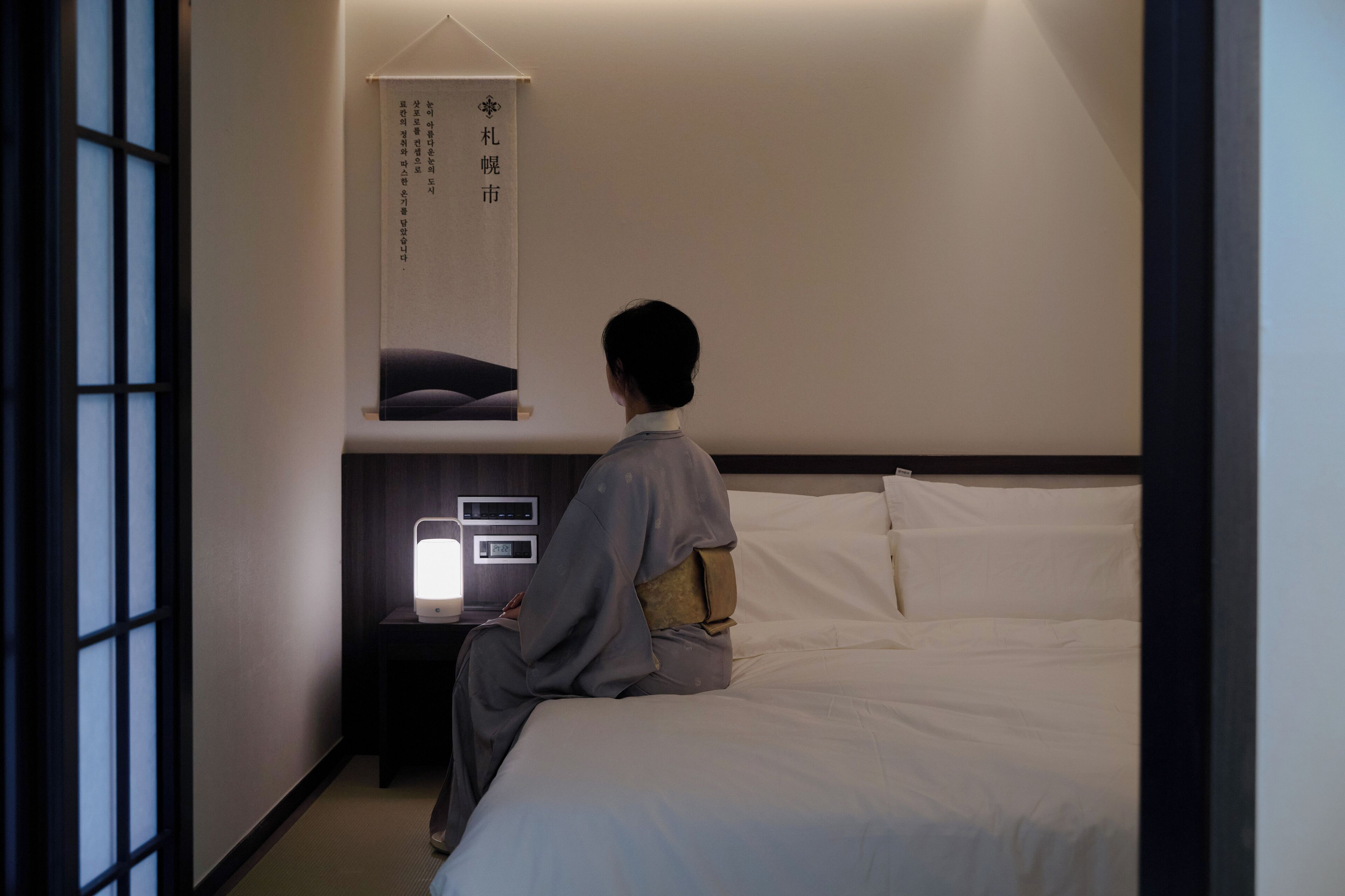 Ein Zimmer mit einem Bett, einem Nachttisch, einem an der Wand befestigten Regal mit einem gerahmten Gegenstand und einem Fenster mit Blick nach draußen.