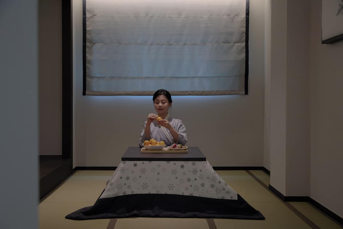 Una persona sentada a una mesa con un plato de comida, en una habitación con suelo de tatami y una mesa baja.