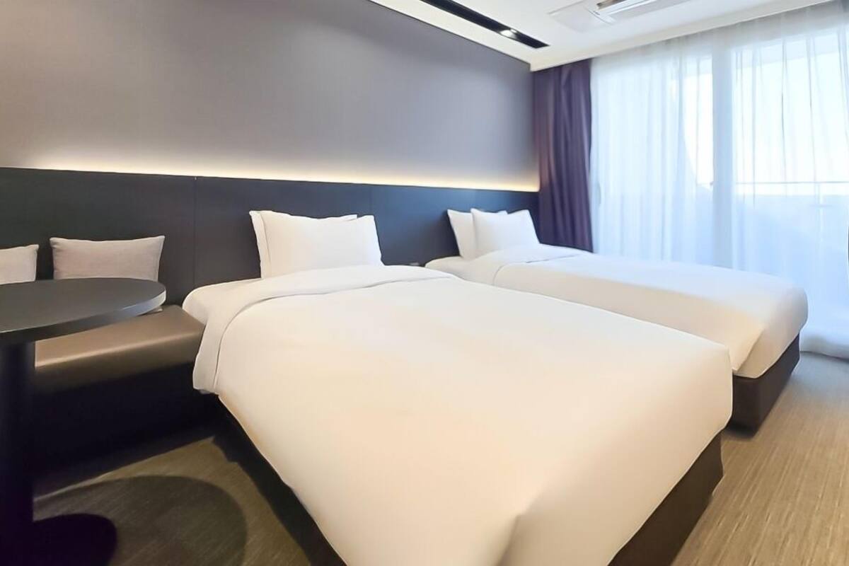 A hotel room with two beds, a small table, and a view of the outdoors through large windows.