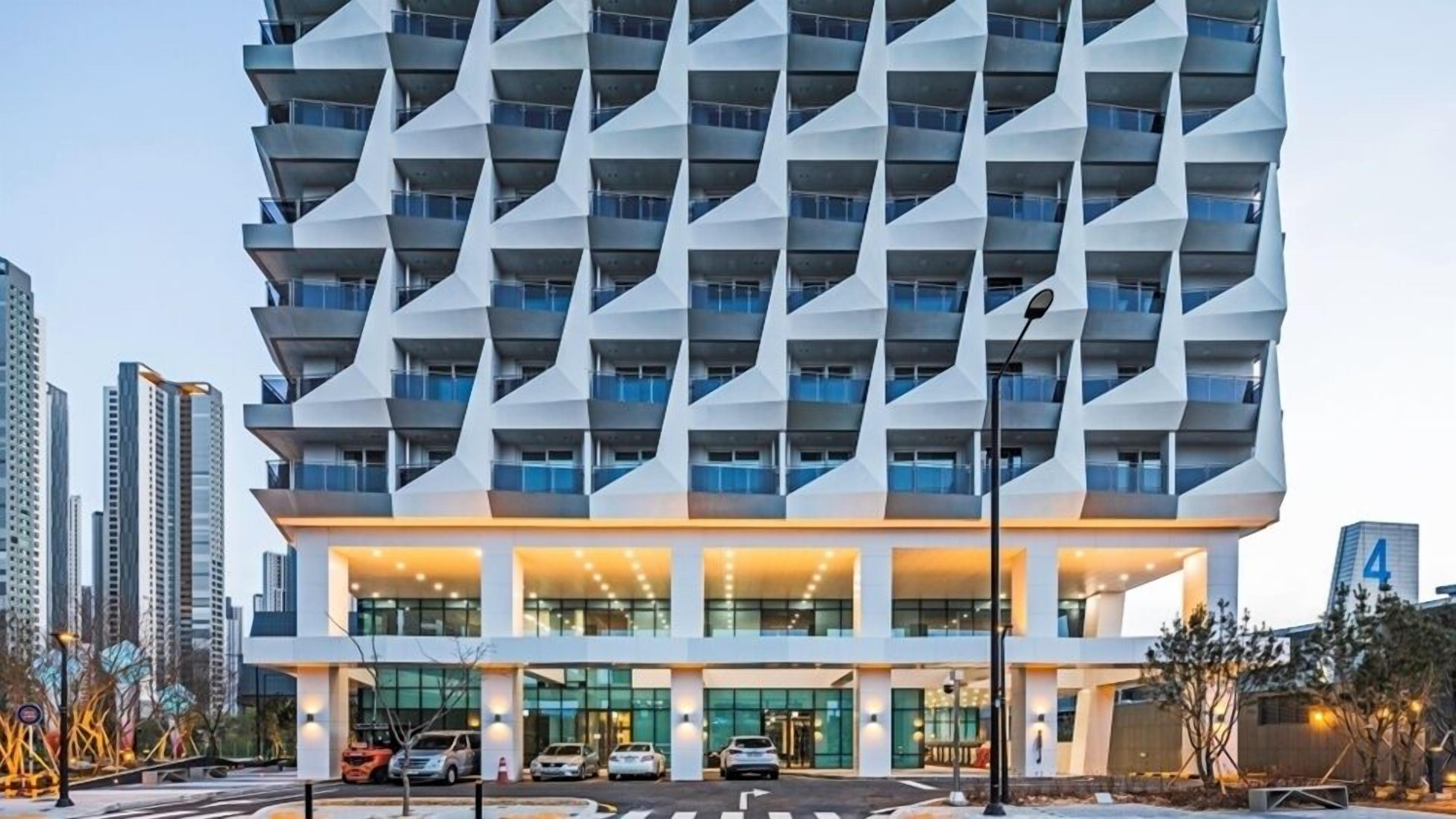 A modern high-rise hotel with a unique geometric facade, multiple balconies, and a well-lit entrance area.
