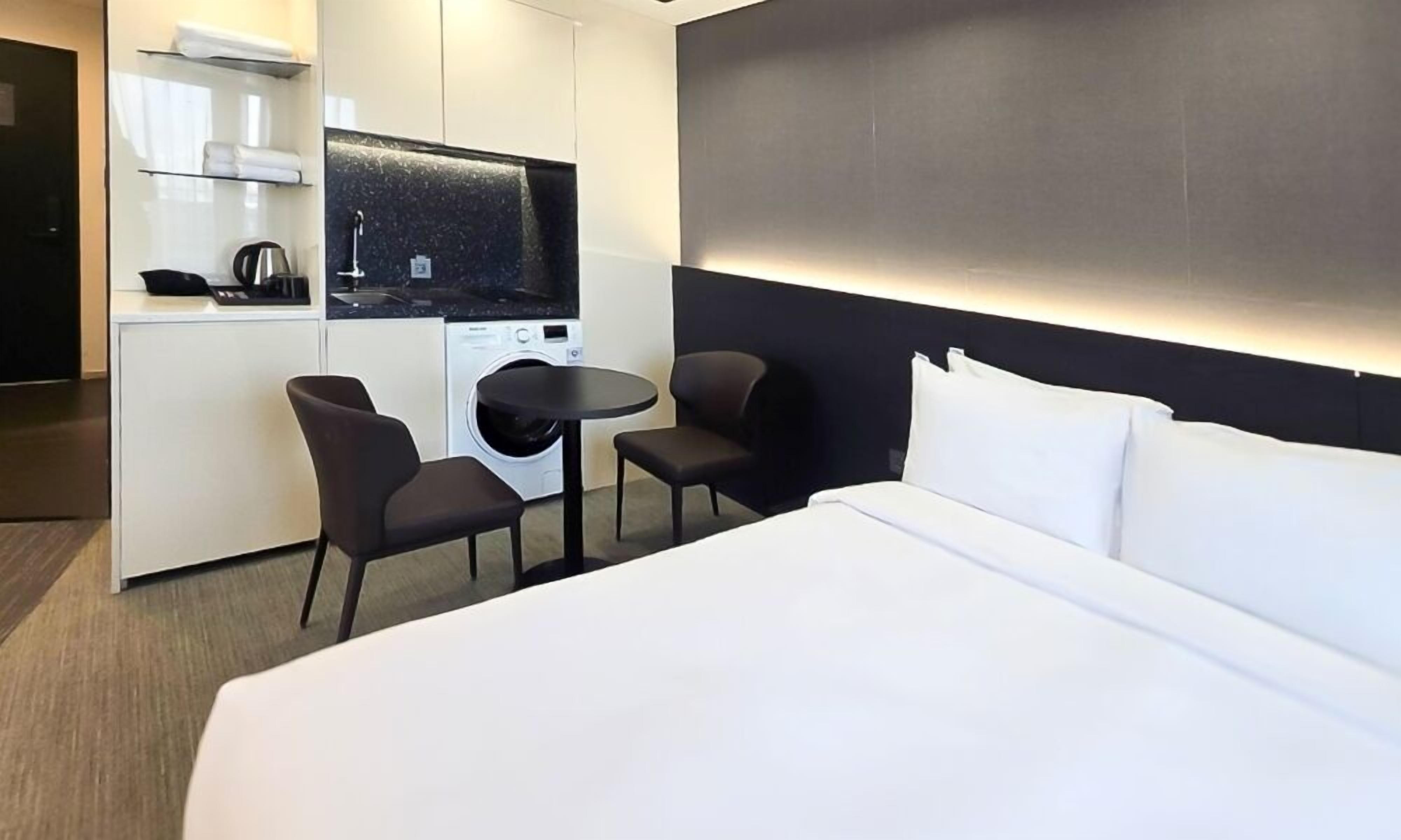 A hotel room with a bed, two chairs, a small table, a washing machine, and a TV.