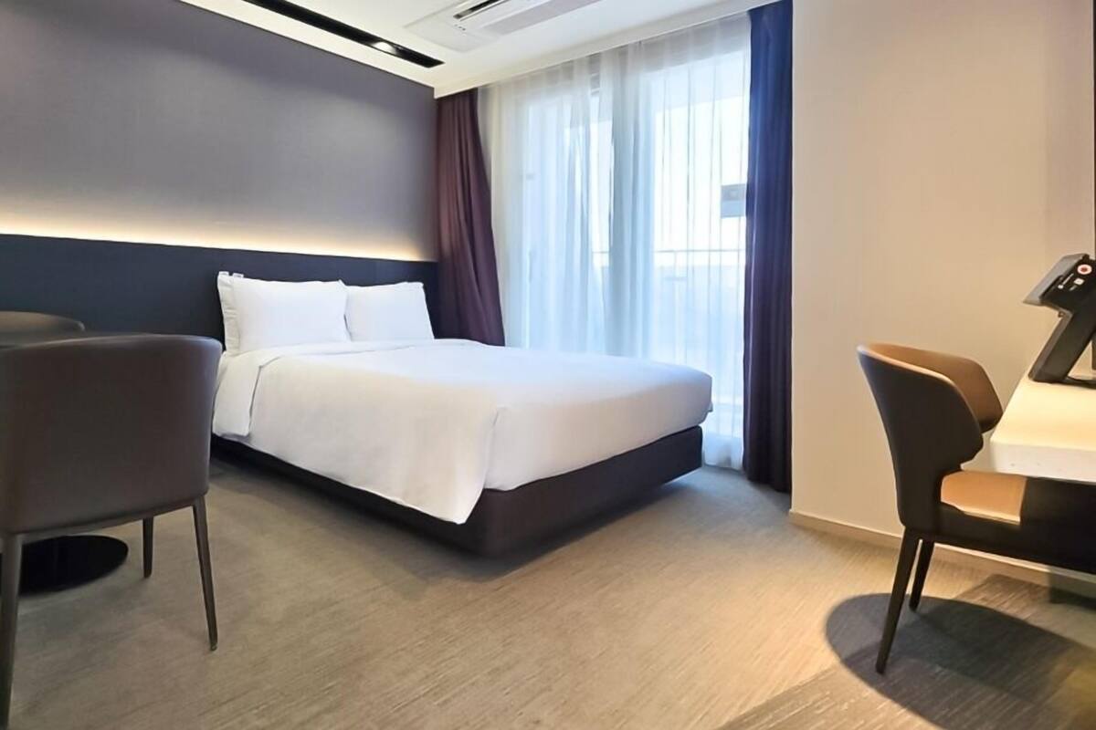 A modern hotel room with a large bed, two chairs, a desk, and a window with curtains.