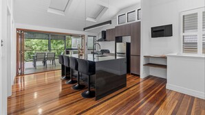 A modern kitchen with a central island, bar stools, and a dining area with a view of greenery. - Elegant Dual-Level Home Near Cafés, Parks & River (East Brisbane)