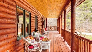 A log cabin with a wooden porch, wicker furniture, and a view of the surrounding trees. - Spacious Log Home w/ 13 Acres, Ponds & Fire Pit! (Kingsley)