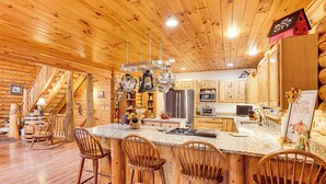 A wooden cabin interior with a kitchen featuring a large island, bar stools, and a staircase leading to another level. - Spacious Log Home w/ 13 Acres, Ponds & Fire Pit! (Kingsley)