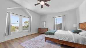 A bedroom with a wooden bed, a nightstand, a dresser, and a ceiling fan. There are two windows with curtains, and a view of the outside is visible. - Luxe Living Suites #3, 3 minutes to DT Cincy (Covington)