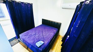 A small, single-bed room with a purple bedspread, a black headboard, and a window with curtains.