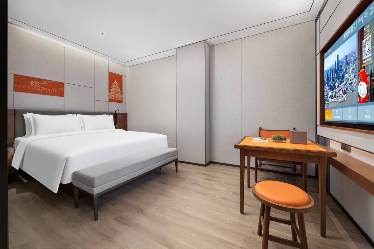 Une chambre d’hôtel moderne équipée d’un lit, d’un bureau avec un ordinateur portable et d’un téléviseur diffusant des images d’une vue urbaine.