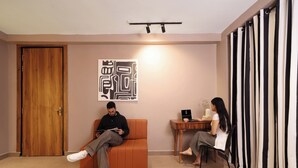 A room with a wooden door, a brown sofa, a small table with a laptop and a vase, and a black and white striped curtain. - The Village Edit - Minimalist Hotels (New Delhi)