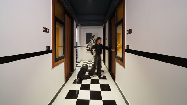 A hotel corridor with checkered flooring, doors numbered 303 and 306, and a person holding a skateboard. - The Village Edit - Minimalist Hotels (New Delhi)
