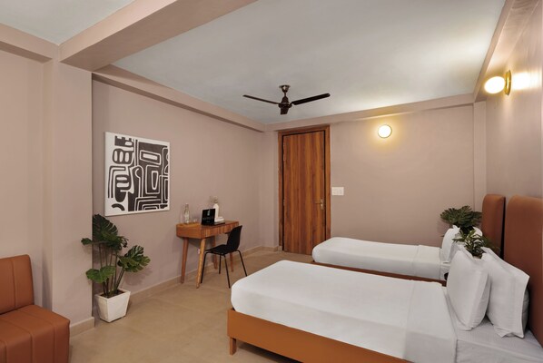 A hotel room with two beds, a desk, a chair, a ceiling fan, and a potted plant. - The Village Edit - Minimalist Hotels (New Delhi)