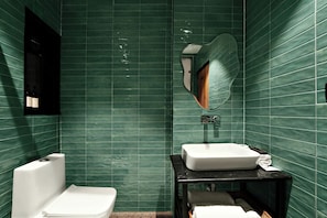 Un baño con paredes de azulejos verdes, un inodoro blanco, un lavabo negro y un espejo con marco de madera.