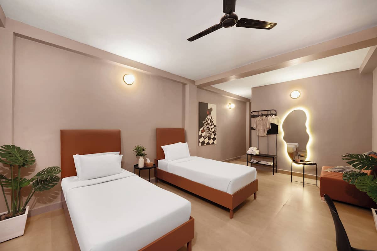 Habitación de hotel moderna con dos camas, ventilador de techo, mesitas de noche, zona de estar y baño con ducha.