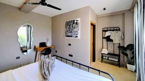 A bedroom with a bed, a desk, a mirror, a wardrobe, and a ceiling fan. - The Village Edit - Minimalist Hotels (New Delhi)