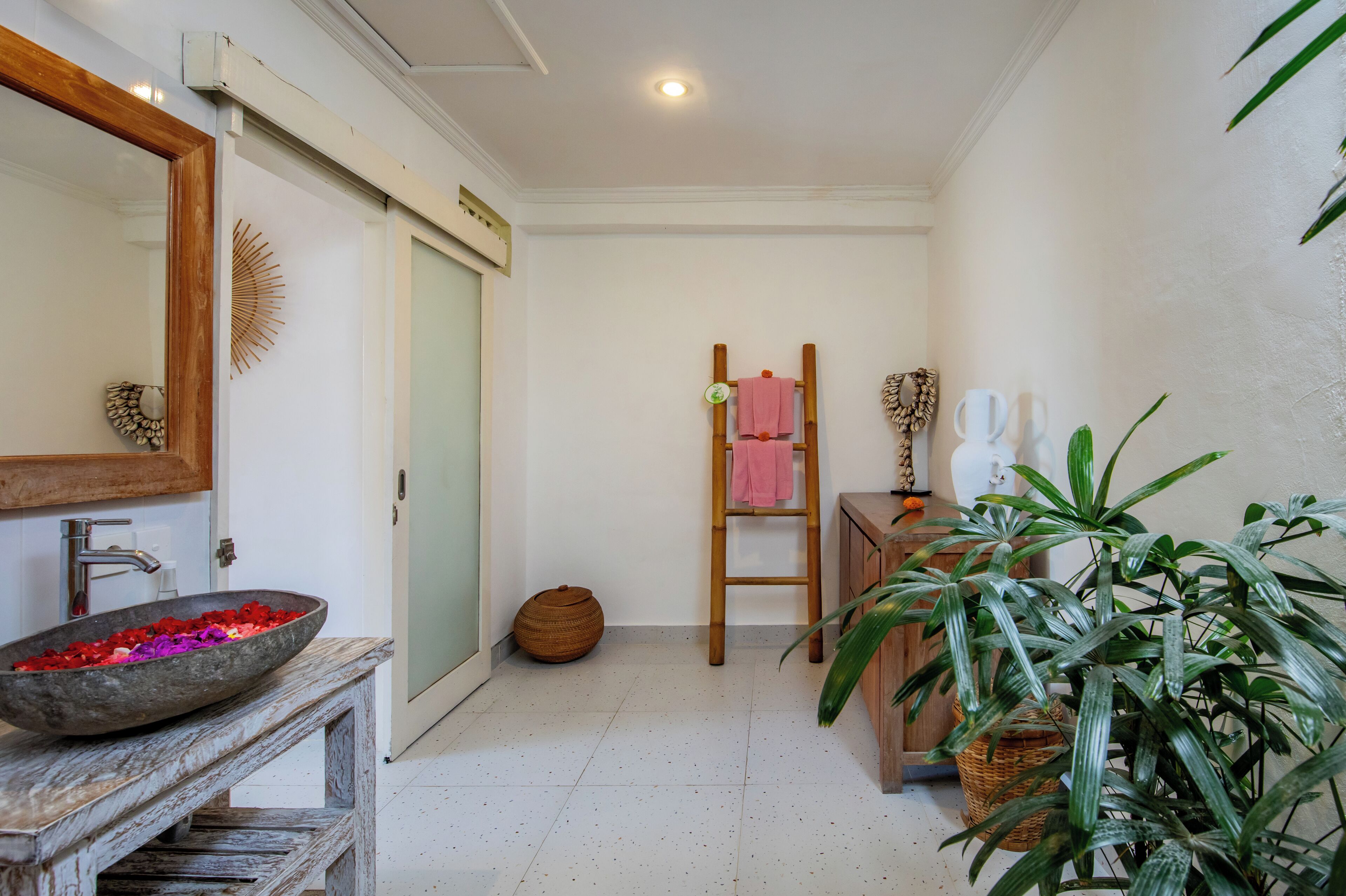 Casa de banho com toucador de madeira, um espelho grande, uma taça com pedras decorativas, uma escada de madeira, uma cesta e uma planta em vaso.