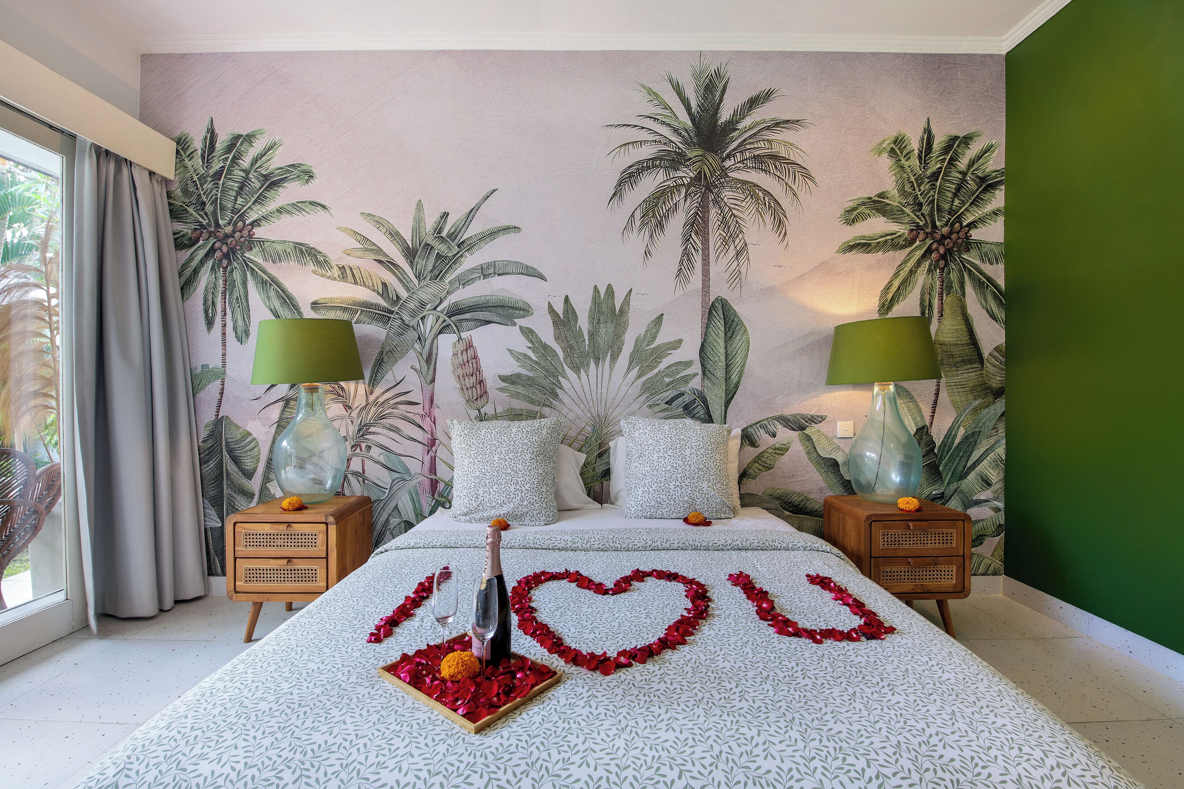 A bedroom with a bed, a mural of palm trees, two nightstands with lamps, and a window with curtains.