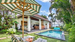 Un espace aménagé au bord de la piscine, comprenant un coin salon abrité, un espace repas et une végétation luxuriante.