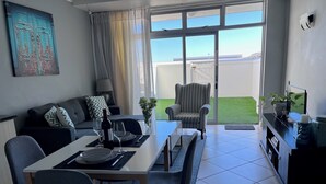A modern living room with a dining area, a sofa, a chair, a table, and a view of a green lawn through large windows. - Stylish 1 Bedroom Big Bay Apartment walking distance from the beach (Cape Town)