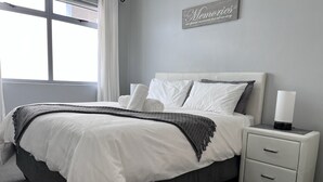 A bedroom with a bed, nightstand, and a wall-mounted sign. - Stylish 1 Bedroom Big Bay Apartment walking distance from the beach (Cape Town)