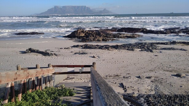 A beach with a wooden walkway leading to the shore, rocky outcrops, and waves crashing onto the sand. - Stylish 1 Bedroom Big Bay Apartment walking distance from the beach (Cape Town)