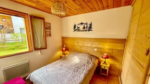 Una camera da letto con soffitto in legno, un letto con copriletto grigio, due comodini con lampade, una finestra vista verde e un muro con la sagoma di una montagna.