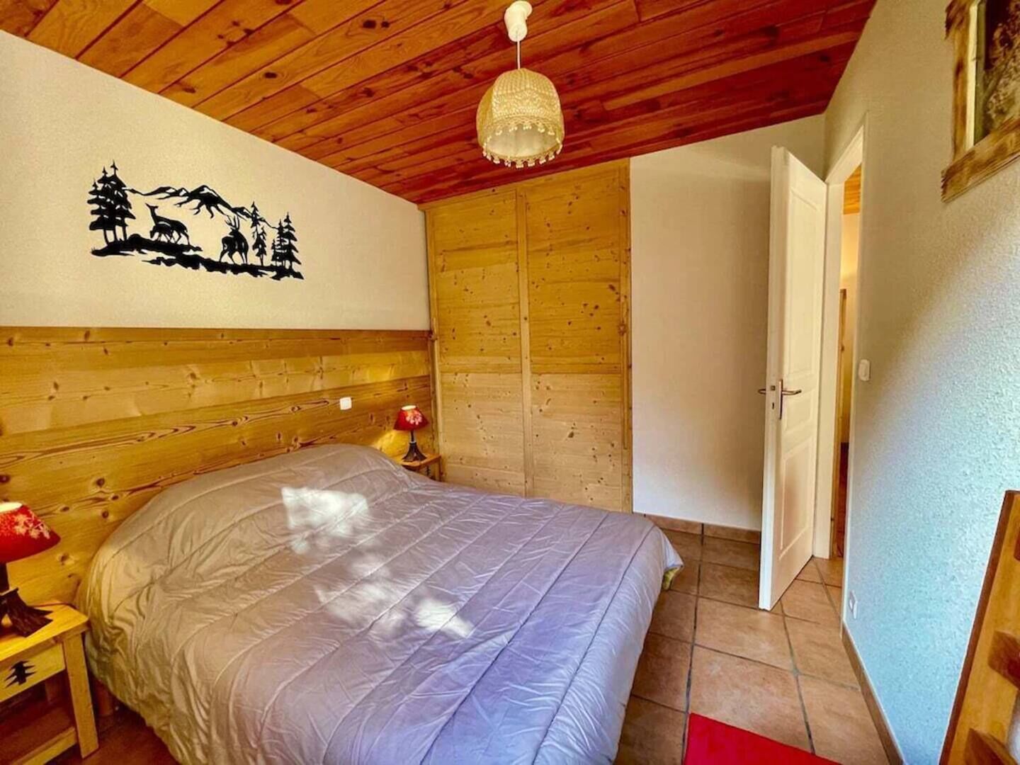 Un dormitorio con techo de madera, una cama con edredón gris, una pared con silueta negra de árboles y animales, y un armario de madera.