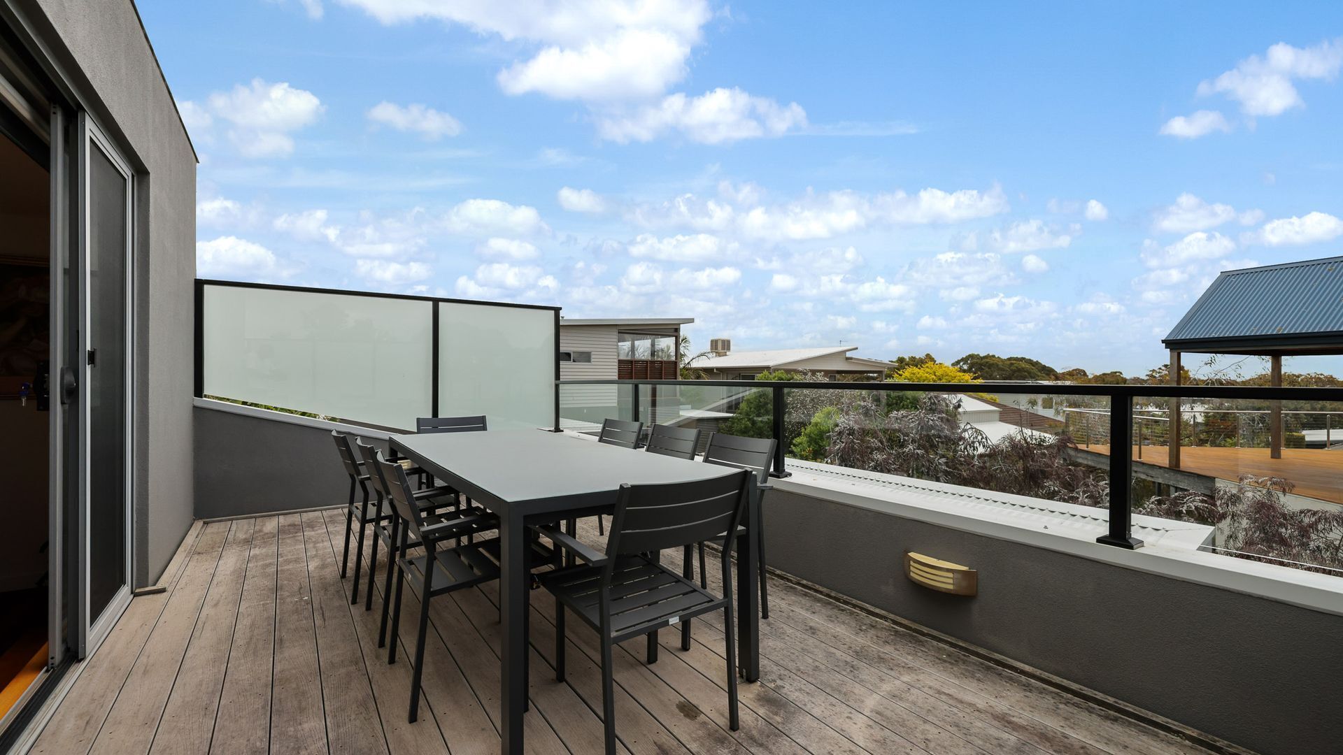 A balcony with a table and chairs, a glass partition, and a view of neighboring buildings and trees.