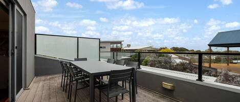 A balcony with a table and chairs, a glass partition, and a view of neighboring buildings and trees.