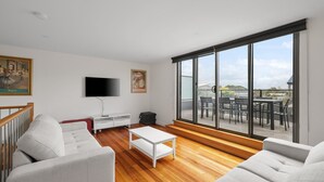Uma sala de estar moderna com uma janela ampla, uma TV de tela plana, um sofá, uma mesa de centro e uma área de jantar com assentos externos.