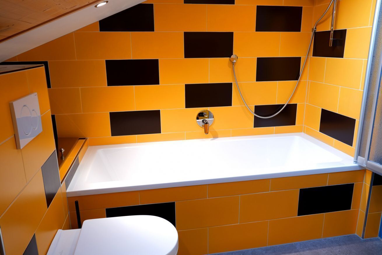 A bathroom with a white bathtub, a shower, and a toilet. The walls are tiled with a pattern of orange and black squares.