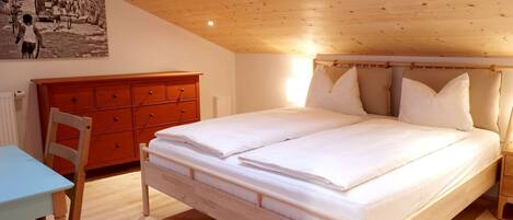 A bedroom with a wooden ceiling, a bed with white bedding, a red dresser, and a small table with a chair.