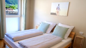 Ein Doppelbett mit zwei Kissen, ein Nachttisch, ein Fenster mit Vorhängen und ein an der Wand hängendes Bild.