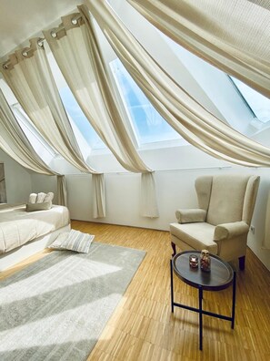 A modern loft bedroom with a large skylight, a comfortable chair, a small table, and a cozy rug. - Comfy 3BR Apt With Living Room Balcony (Vienna)