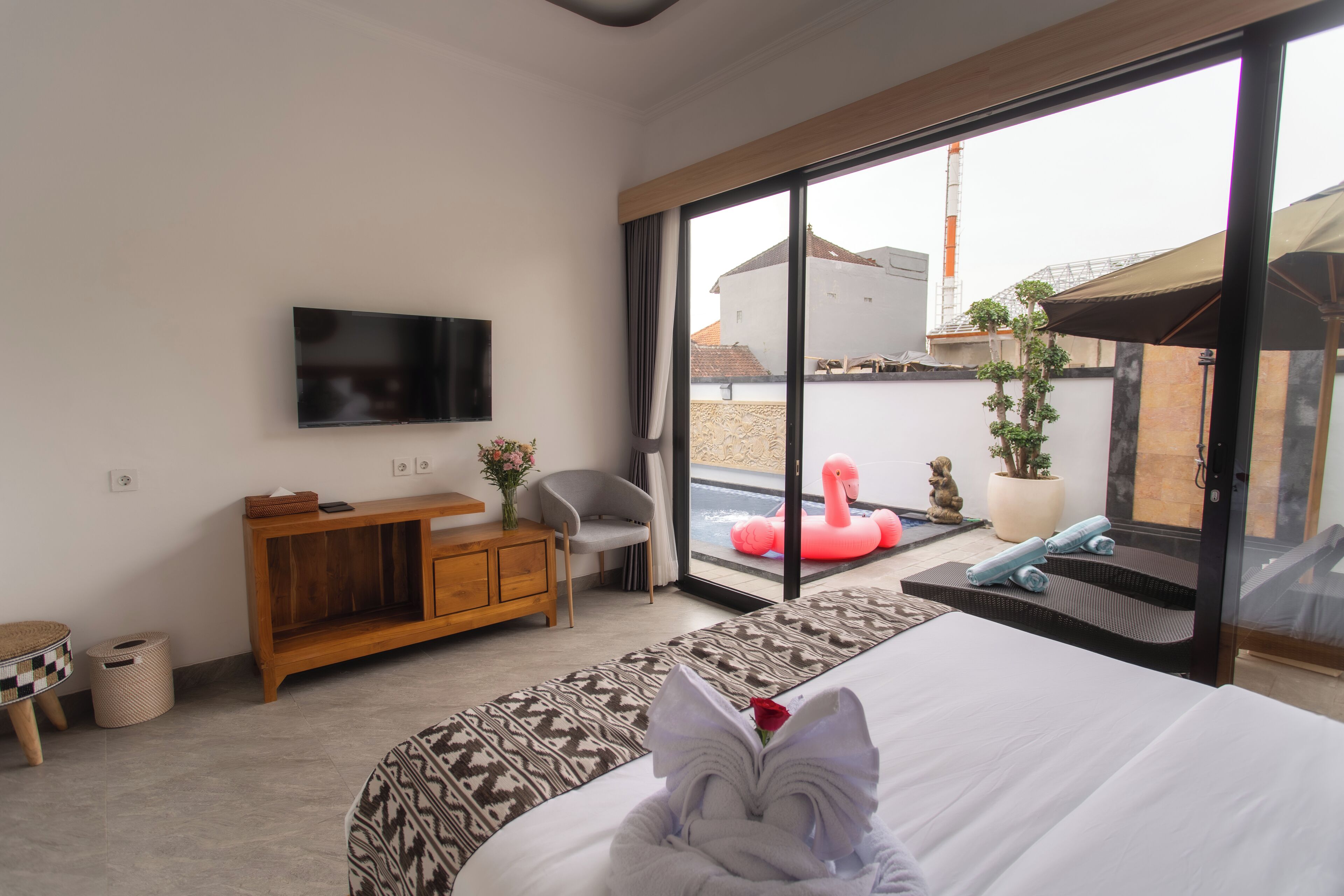 A modern hotel room with a large bed, a TV mounted on the wall, a wooden side table, a chair, and a view of a swimming pool and outdoor area.